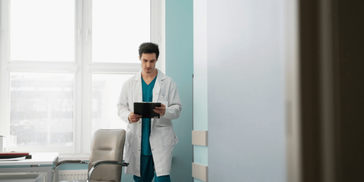 Doctor in a white coat standing in a medical office, reading a tablet.