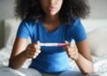 Disappointed hispanic girl getting unexpected result from pregnancy test kit. Sad young latina woman sitting alone on her bed.