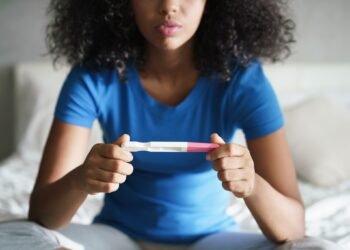 Disappointed hispanic girl getting unexpected result from pregnancy test kit. Sad young latina woman sitting alone on her bed.