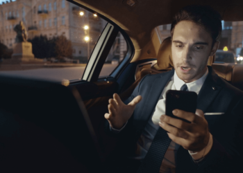 A man in a suit and tie sits in a car's back seat, focused on his cell phone.