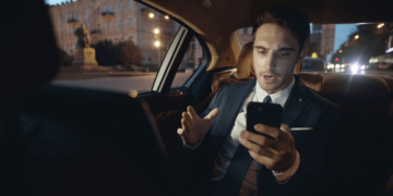 A man in a suit and tie sits in a car's back seat, focused on his cell phone.