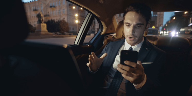 A man in a suit and tie sits in a car's back seat, focused on his cell phone.