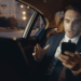 A man in a suit and tie sits in a car's back seat, focused on his cell phone.