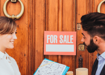 A man and woman stand together in front of a door, holding a for sale sign, indicating a property listing.