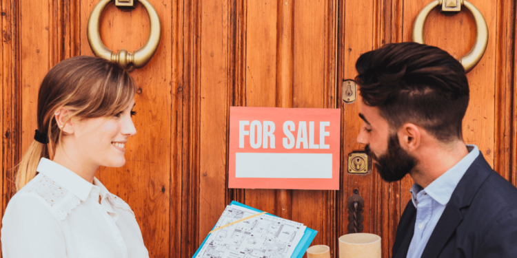 A man and woman stand together in front of a door, holding a for sale sign, indicating a property listing.