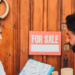 A man and woman stand together in front of a door, holding a for sale sign, indicating a property listing.