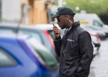 Young Male Security Guard In Black Uniform Using Walkie-Talkie On Street