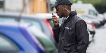 Young Male Security Guard In Black Uniform Using Walkie-Talkie On Street