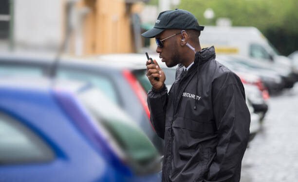 Young Male Security Guard In Black Uniform Using Walkie-Talkie On Street